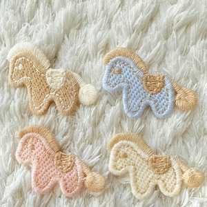 Puede incluir: Cuatro parches bordados de caballo con un diseño estilizado que incluye crin, cola y silla de montar. Los parches son de color beige, azul claro, rosa suave y crema, cada uno con crin y cola beige, mostrando un trabajo de hilo detallado.