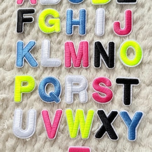 Può includere: Lettere dell'alfabeto ricamate in vari colori, tra cui rosa, blu, giallo e nero, con bordi bianchi. Le lettere sono disposte su una superficie bianca e strutturata, creando un'esposizione colorata e giocosa.