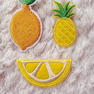 Może przedstawiać: Trzy haftowane naszywki: żółta cytryna, żółty ananas i żółta plaster cytryny. Cytryna i ananas mają zielone akcenty. Naszywki znajdują się na białym, puszystym tle.