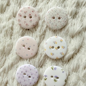 Può includere: Sei bottoni rotondi con motivi floreali nei toni del rosa, bianco e viola. Ogni bottone ha due fori per cucire. I bottoni sono disposti su una superficie morbida e strutturata di colore bianco.