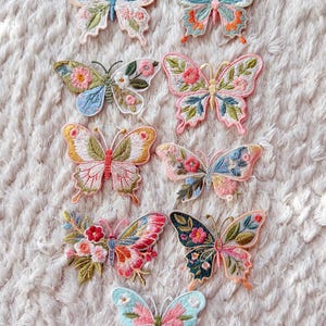 Op de afbeelding: Een verzameling van negen geborduurde vlinderpatches met ingewikkelde bloemen- en botanische patronen in diverse kleurenpaletten, waaronder roze, blauw, groen, oranje en geel. Elke patch toont gedetailleerd borduurwerk en unieke vleugelvormen.