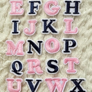 May include: Embroidered alphabet patches in pink and navy blue. Each letter has a pink center and a navy blue outline, with a white border. The letters are arranged alphabetically on a textured white surface.