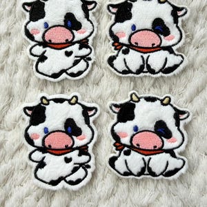 May include: Four embroidered patches featuring a cartoon cow design. Each patch has a white body with black spots, a pink nose, blue eyes, and a red scarf. The patches are on a white, textured background.