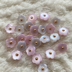 以下が含まれることがあります： 白いふわふわの表面に、金色のセンターを持つピンクと白の花の形をしたボタンが20個集められています。ボタンはランダムなパターンで配置されています。