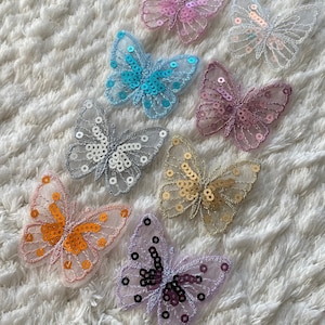 Könnte beinhalten: Sortiment von acht Schmetterlings-Applikationen aus Stoff mit Pailletten. Die Schmetterlinge sind in verschiedenen Farben, darunter rosa, blau, weiß, gold, orange und lila.