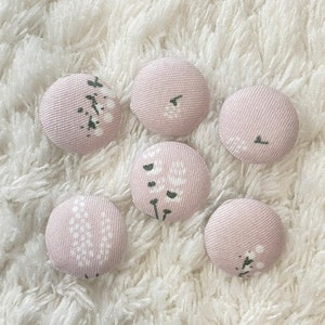 May include: Six round, fabric-covered buttons in a soft pink hue. Each button features a delicate white floral design with green accents. The buttons are arranged on a fluffy, white textured surface.