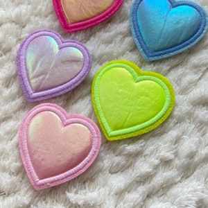 May include: Five heart-shaped patches with iridescent fabric and contrasting colored stitching. The patches are pink, purple, blue, green, and yellow.