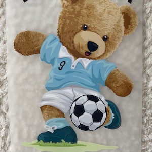 May include: A brown teddy bear wearing a blue soccer jersey with the number 9 on it, kicking a black and white soccer ball. The text "TOP SCORER" is written in black above the bear.