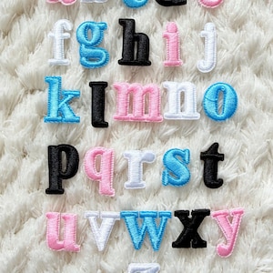 May include: Embroidered lowercase alphabet letters in various colors, including pink, blue, black, and white. The letters are arranged on a white, fluffy surface, ideal for crafting or personalization projects.