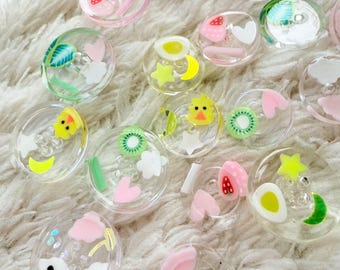 May include: A collection of clear, round resin cabochons with various embedded designs. The designs include fruits, stars, clouds, and smiling faces in shades of pink, yellow, green, and white. The cabochons are scattered on a fluffy, white surface.