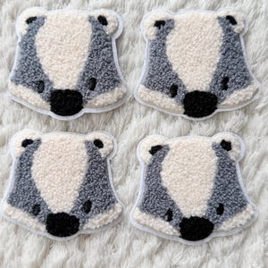 Könnte beinhalten: Vier fuchsähnliche Aufnäher mit flauschiger, strukturierter Optik. Jeder Aufnäher hat ein weißes Gesicht, einen grau-weißen Körper sowie eine schwarze Nase und Augen. Die Aufnäher sind vor einem weißen, flauschigen Hintergrund platziert.