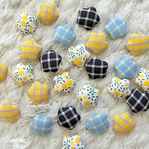 May include: A collection of small, star-shaped fabric buttons featuring various patterns including yellow and blue plaid, black and white gingham, and white fabric with a yellow and green lemon print scattered across the surface.