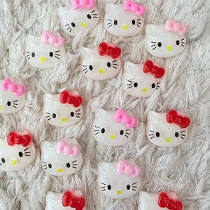 Może przedstawiać: Kolekcja białych, błyszczących zawieszek z twarzą Hello Kitty z różowymi i czerwonymi kokardkami. Każda zawieszka ma czarne wąsy i oczy oraz żółty nos. Zawieszki są ułożone na białej, teksturowanej powierzchni.