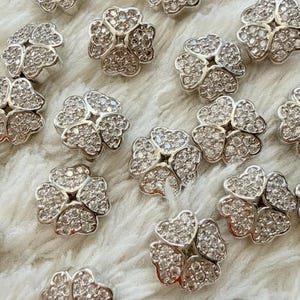 Può includere: Una collezione di bottoni in metallo color argento, ognuno a forma di quadrifoglio. La superficie di ogni bottone è densamente ricoperta da piccole pietre chiare e riflettenti che creano un aspetto scintillante e strutturato.