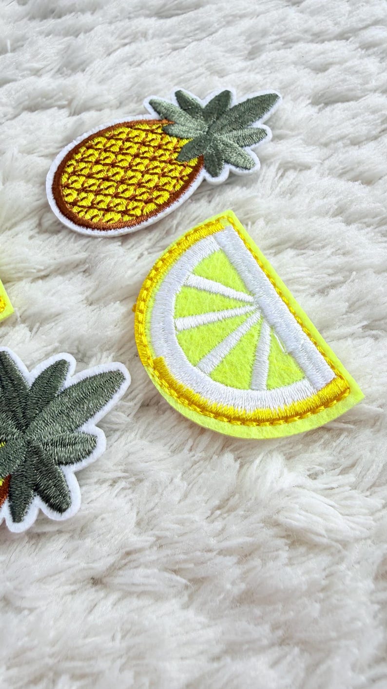 May include: Embroidered patches of a pineapple and a lemon slice. The pineapple is yellow with a brown border and green leaves. The lemon slice is yellow with a white rind and yellow stitching. These patches are likely for clothing or accessories.