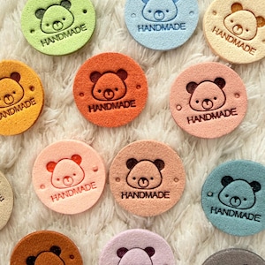 Op de afbeelding: Een verzameling kleurrijke, ronde "HANDMADE" labels met een berengezicht ontwerp. De labels zijn in verschillende pastelkleuren, waaronder groen, blauw, perzik en bruin. De labels zijn gemaakt van een zacht, getextureerd materiaal en liggen op een witte, pluizige ondergrond.