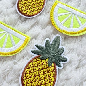 May include: Embroidered patches of a pineapple and lemon slices. The pineapple patches have a yellow body with brown outlines and green leaves. The lemon slices are yellow with white outlines. These patches are ideal for adding a touch of summer to clothing or accessories.