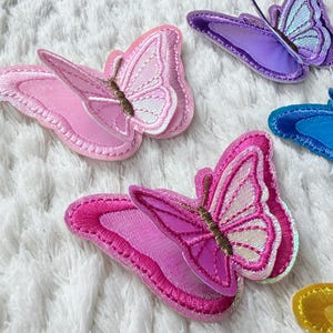 May include: A collection of colorful butterfly patches in shades of pink, purple, and blue. Each patch features detailed embroidery and a layered design, with a brown antenna. The patches are arranged on a white, textured surface.