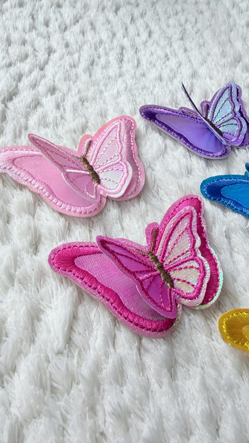 May include: A collection of colorful butterfly patches in shades of pink, purple, and blue. Each patch features detailed embroidery and a layered design, with a brown antenna. The patches are arranged on a white, textured surface.