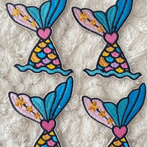 May include: Four embroidered iron-on patches featuring a mermaid tail with a pink heart, yellow, blue, and purple scales, and gold stars on a pink background. The tail is blue and turquoise with a white outline.