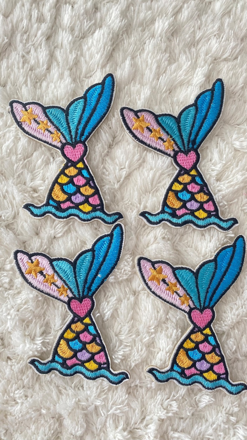 May include: Four embroidered iron-on patches featuring a mermaid tail with a pink heart, yellow, blue, and purple scales, and gold stars on a pink background. The tail is blue and turquoise with a white outline.