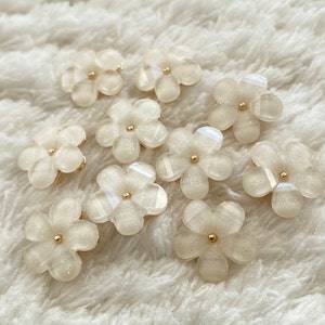 May include: Twelve white glitter flower embellishments with gold centers. The flowers are scattered on a white fuzzy surface.