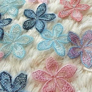 Può includere: Una collezione di toppe ricamate a forma di fiore in vari colori, tra cui azzurro, blu scuro, rosa e viola. Ogni toppa presenta cinque petali con un centro in rete trasparente e cuciture dettagliate lungo i bordi dei petali.