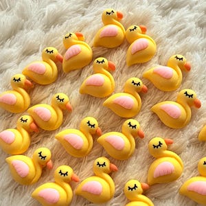 Peut inclure: Plusieurs figurines de canards jaunes avec des ailes roses et des becs oranges. Les canards ont des yeux noirs et sont disposés sur une surface blanche et moelleuse. Ces figurines décoratives sont idéales pour l'artisanat ou comme charmants accents.