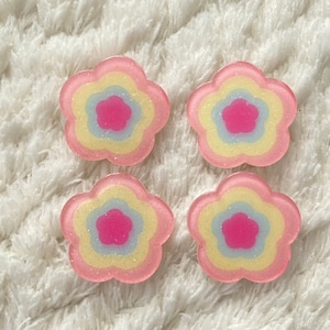 Puede incluir: Cuatro adornos de resina en forma de flor rosa con círculos concéntricos amarillos y azules. Los adornos tienen un acabado brillante.