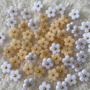 Può includere: Una collezione di bottoni a forma di fiore di colore bianco e crema con centri dorati. I bottoni sono disposti in modo casuale su una superficie bianca e pelosa.