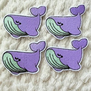 Pode incluir: Quatro patches bordados com um design de baleia de desenho animado. As baleias são roxas com barrigas verde claro e uma borda branca. Os patches estão em uma superfície branca texturizada.