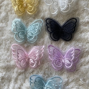 Könnte beinhalten: Sieben gestickte Schmetterlingsapplikationen in verschiedenen Farben, darunter Gelb, Weiß, Blau, Schwarz, Rosa, Lila und Hellblau. Die Schmetterlinge sind auf einer weißen, flauschigen Oberfläche übereinander gelegt.