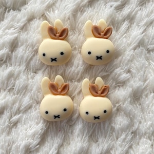 May include: Four white bunny-shaped buttons with brown ears and black eyes. The buttons are on a white fuzzy surface.