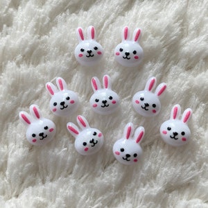 May include: Nine white plastic bunny buttons with pink ears and black eyes. The buttons are arranged in a semi-circle on a white fluffy surface.