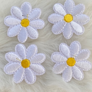 May include: Four white embroidered daisy patches with yellow centers. The patches are on a white fluffy surface.