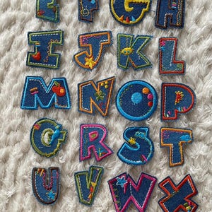 Könnte beinhalten: Ein Set mit 26 Buchstaben-Patches aus Denim mit bunten Stickereien. Jeder Buchstabe ist mit verschiedenen Designs verziert, darunter Sterne, Blumen und geometrische Formen.