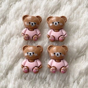 Op de afbeelding: Vier bruine teddybeer-knopen met roze kleding en roze voeten. De knopen liggen op een wit, pluche oppervlak.