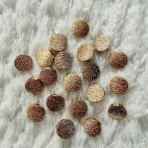 Puede incluir: Una colección de pequeños botones redondos de color dorado con una superficie texturizada. Los botones están dispersos sobre un fondo blanco, esponjoso y texturizado. Los botones parecen ser de metal.