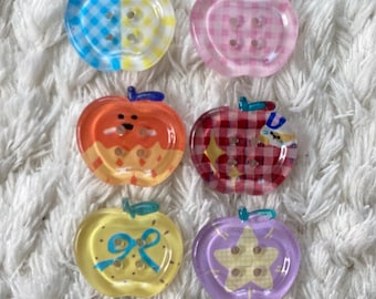 May include: Six colorful apple-shaped buttons with various patterns and designs. The buttons feature a mix of colors, including blue, pink, orange, red, yellow, and purple. Each button has a stem and is set against a white, textured background.