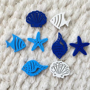 Puede incluir: Una colección de recortes de madera con temática marina, que incluye peces, estrellas de mar y conchas. Los recortes son en tonos azules y blancos, dispuestos sobre una superficie blanca texturizada. Las formas son detalladas y decorativas.