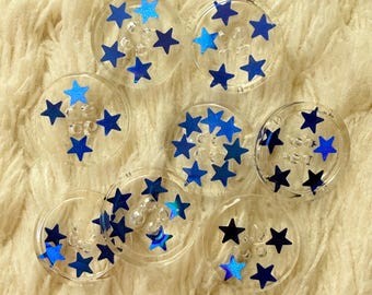 May include: Clear, circular embellishments with blue star designs. The stars vary in shades of blue, including a shimmering, iridescent effect. The embellishments are arranged on a soft, cream-colored, textured surface.