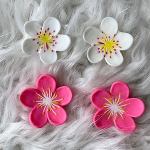 May include: Four decorative flower embellishments, two white and two pink, with yellow centers and white accents. The flowers are on a white fluffy surface.