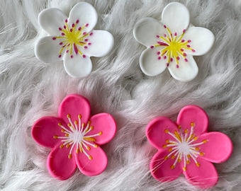 May include: Four decorative flower embellishments, two white and two pink, with yellow centers and white accents. The flowers are on a white fluffy surface.