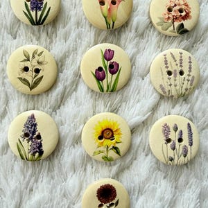 Può includere: Una collezione di bottoni in legno, ognuno con un diverso design floreale. I bottoni presentano vari fiori, tra cui calle, ortensie, tulipani, lavanda e un girasole. I bottoni sono rotondi e hanno due fori per cucire.