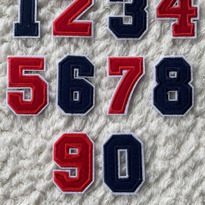 Può includere: Dieci toppe da stirare con i numeri da 1 a 10. I numeri sono in uno stile di font universitario. I numeri 1, 3, 6, 8 e 9 sono blu navy con contorni bianchi. I numeri 2, 4, 5, 7 e 10 sono rossi con contorni bianchi.
