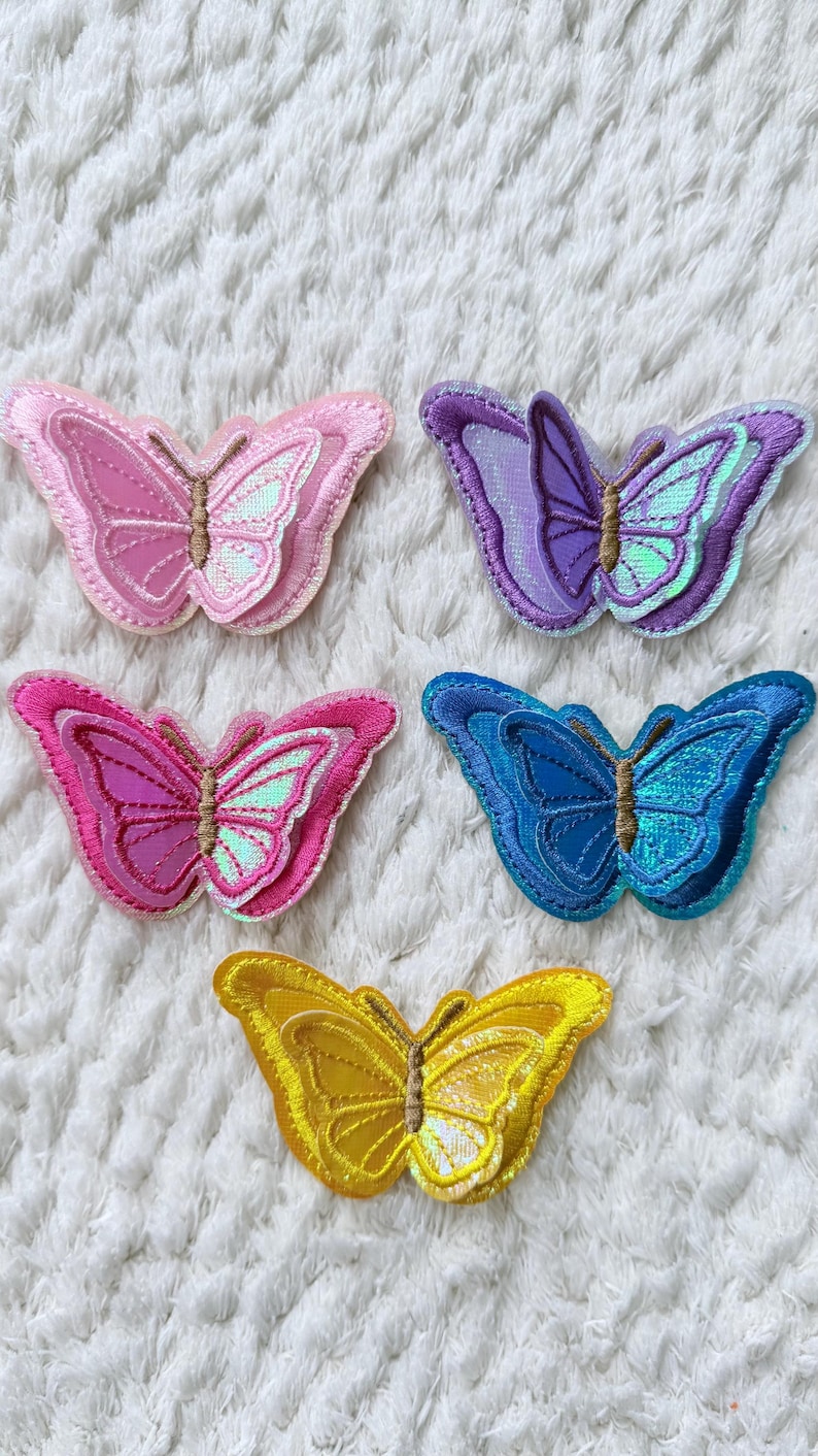 May include: Five embroidered butterfly patches in shades of pink, purple, blue, and yellow. Each butterfly has a layered design with iridescent accents. The patches are suitable for clothing or craft projects.