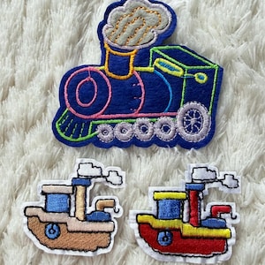 May include: Three colourful embroidered patches: a blue train with pink and green accents, and two small boats. The train has a smoke cloud and detailed wheels. The boats are tan and red with blue and yellow details.