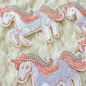 Può includere: Molteplici toppe ricamate a forma di unicorno con corpo bianco, criniera verde menta chiaro e rosa corallo, e un corno abbinato color rosa corallo. Ogni toppa raffigura un unicorno al galoppo con cuciture dettagliate e un bordo bianco.