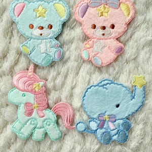 May include: Four embroidered patches in pastel colours: two teddy bears, a unicorn, and an elephant. The bears are light blue and pink, the unicorn is mint green, and the elephant is blue. Each has a star and bow.