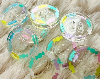 May include: Clear, round buttons with colorful candy designs. Each button features a ring of pastel-colored candies in shades of blue, green, yellow, and pink. The buttons have four holes for sewing and are set against a soft, white background.